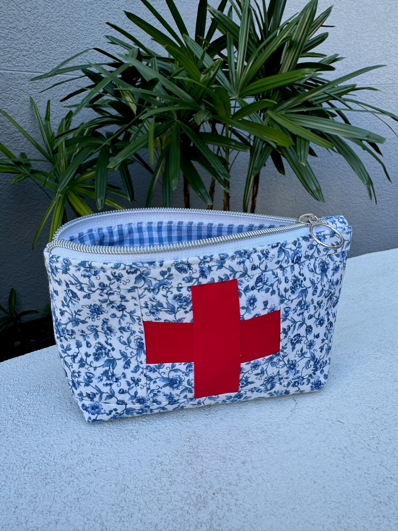 Floral-patterned pouch with a red cross on a white surface, plant in the background