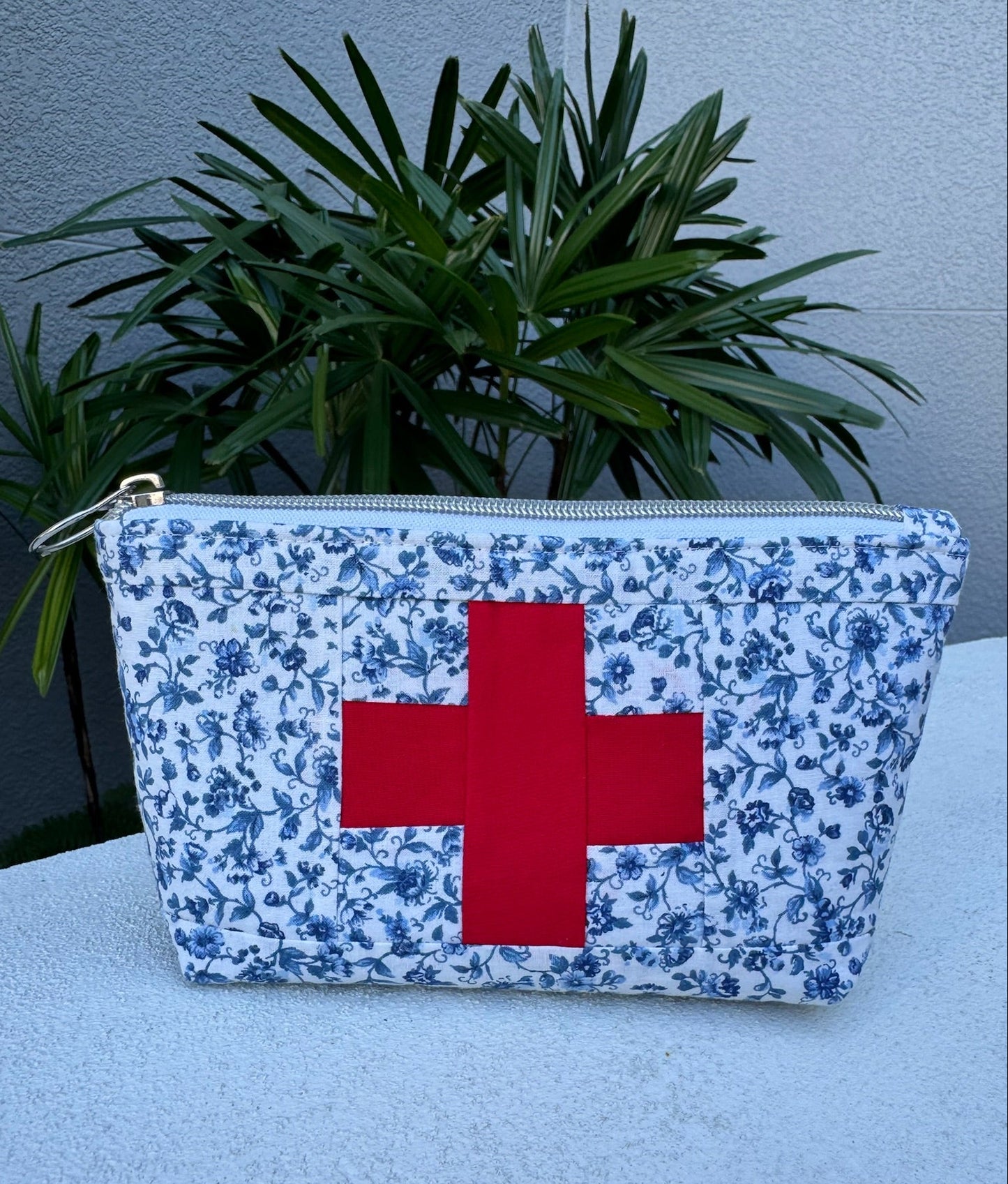 First aid kit pouch with red cross on blue floral pattern, placed on a light surface with a plant in the background.